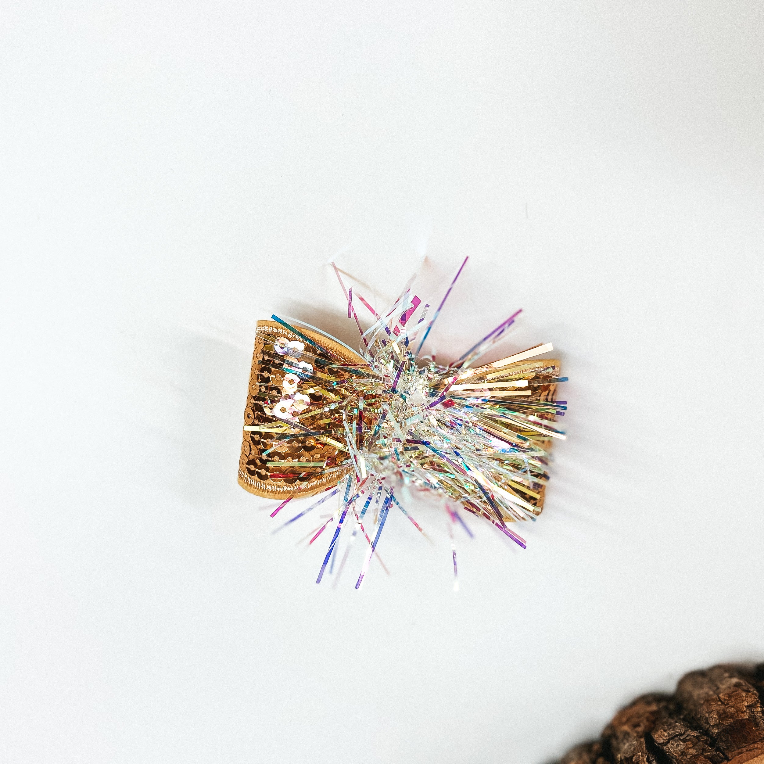 This is a small gold sequin bow with a tinsel center, this bow is taken on a  white background with a slab of wood in the corner as decor.