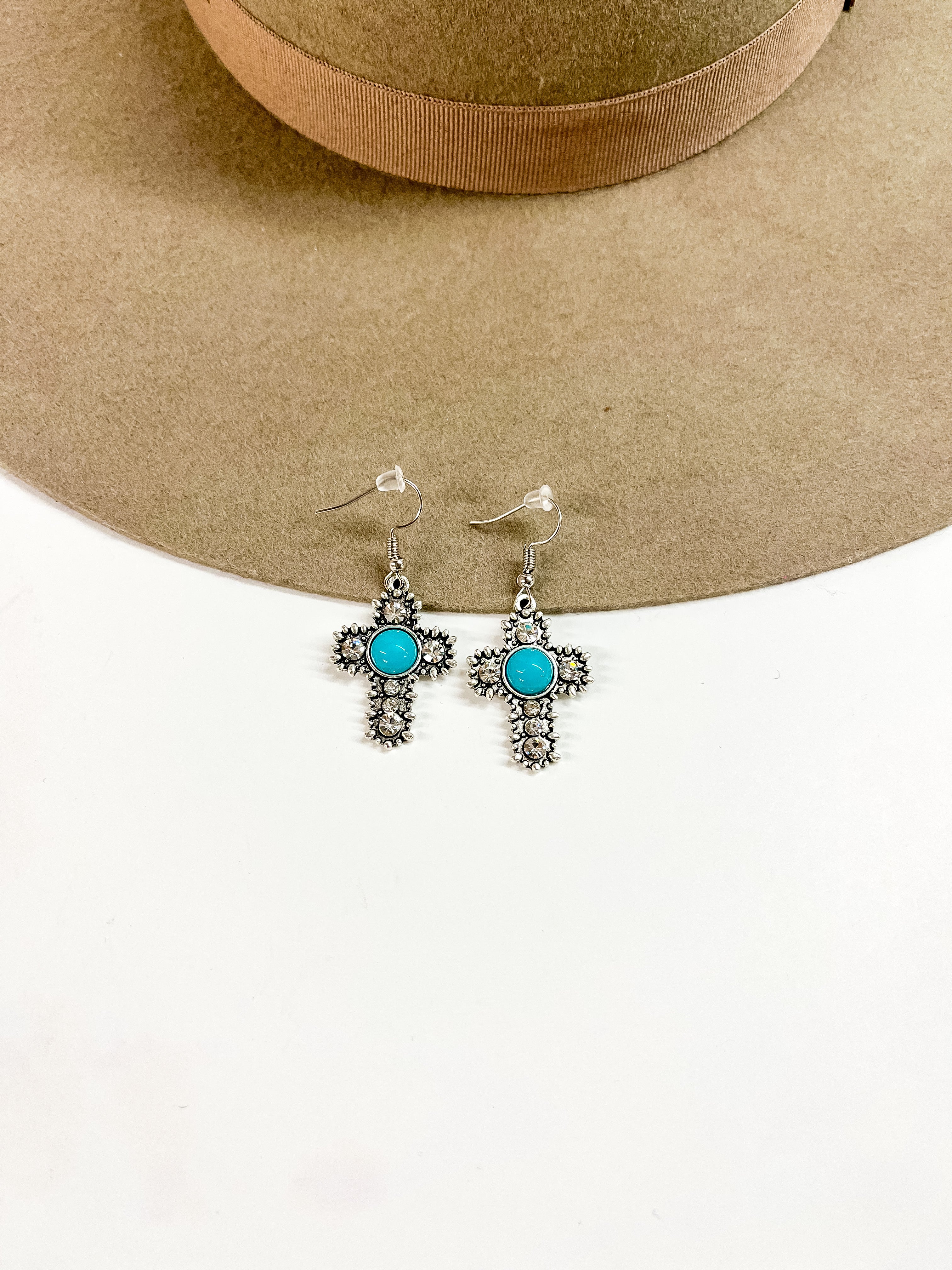 This is a pair of silver detailed cross earrings with small clear crystals  and a turquoise circle stone in the center.  These earrings are laying on a brown felt hat brim and on a white background.