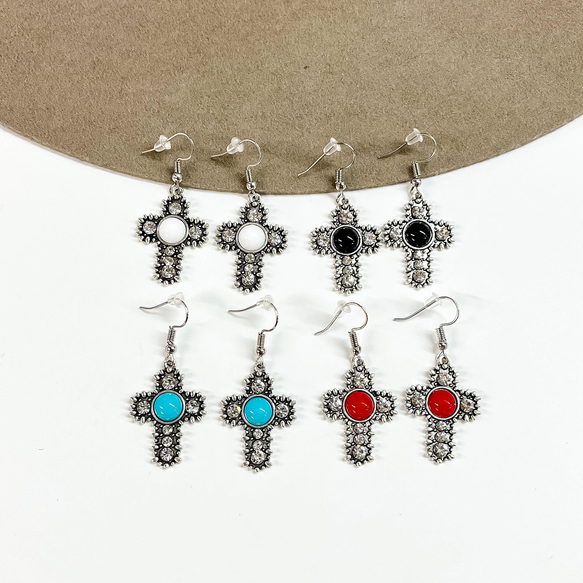 There are four pairs of silver detailed cross earrings with different colored stones in the center. From top to bottom; white, black, turquoise, and red. These earrings are taken partly laying on a brown felt hat brim and on white background.