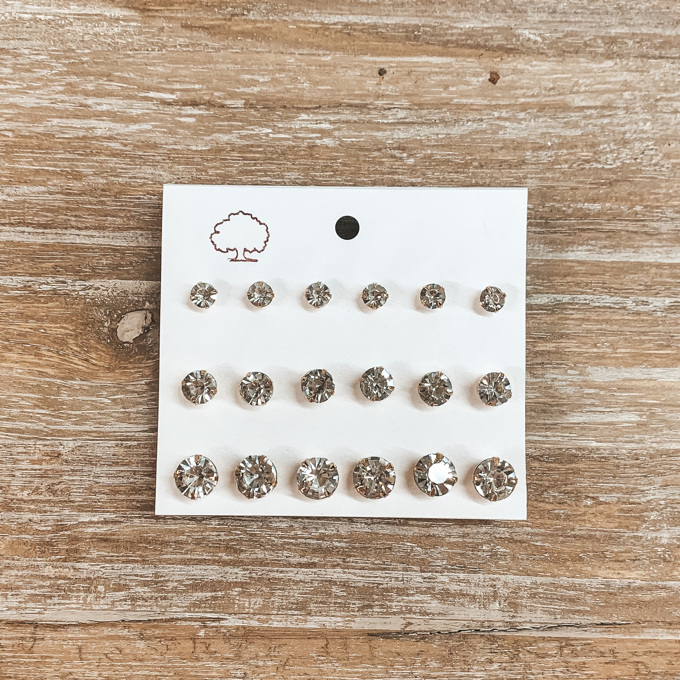 This is a pack of nine crystal clear studs in a gold settings in different sizes. These earrings are placed on a white earring holder and taken on a wooden slate.