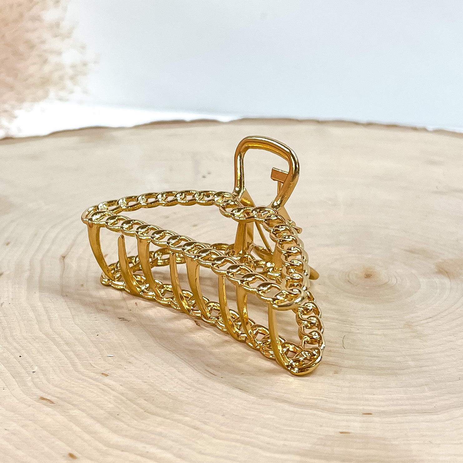 This is a small dome shaped hair clip with chain texture in gold. This hair  clip is laying on a slab of wood with a beige plant in the side as decor.