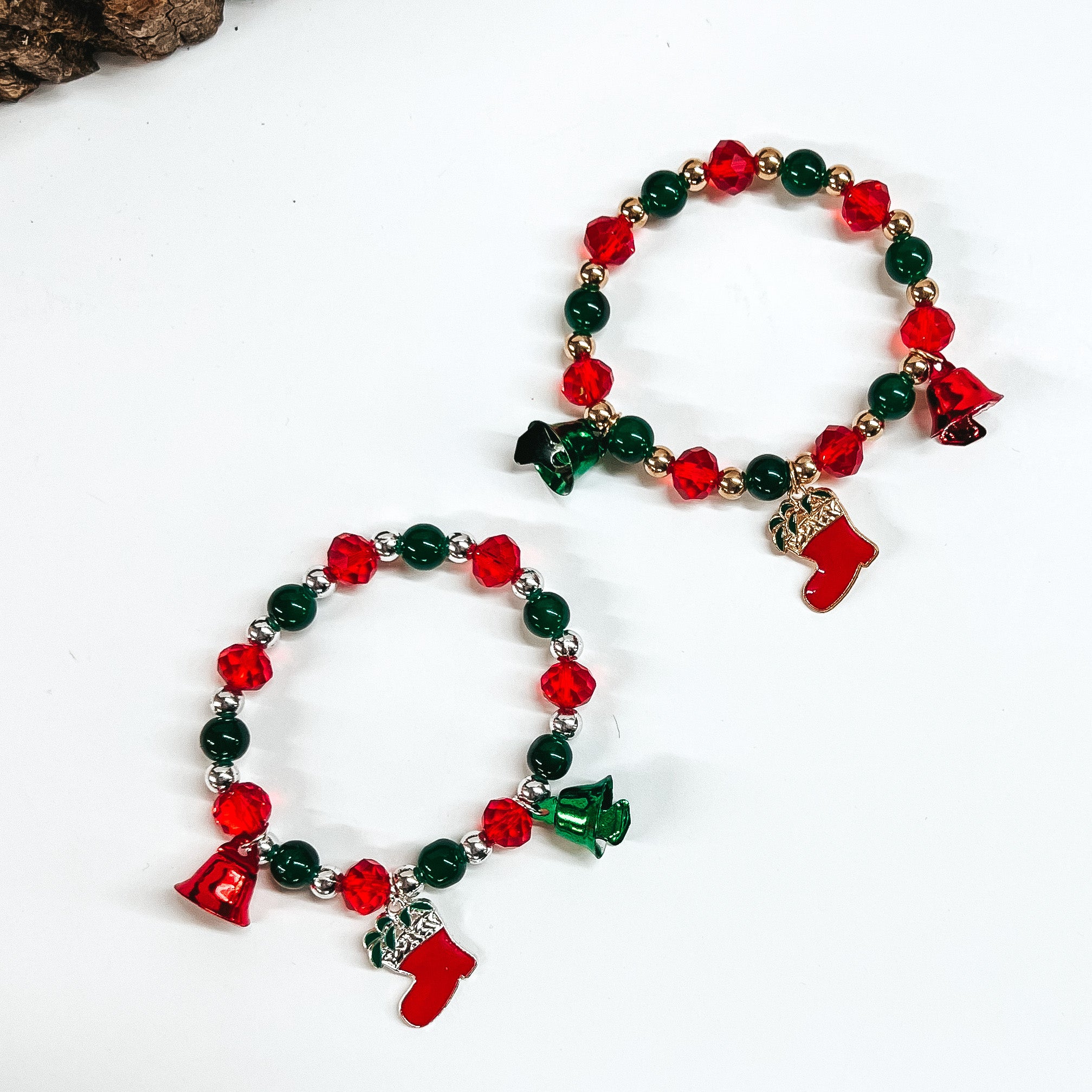 There are two red and green beaded bracelets with charms. The bottom one has silver circle beads and the top on ehas gold circle beads. Both bracelets are taken on a white background with a slab of wood in the back as decor.