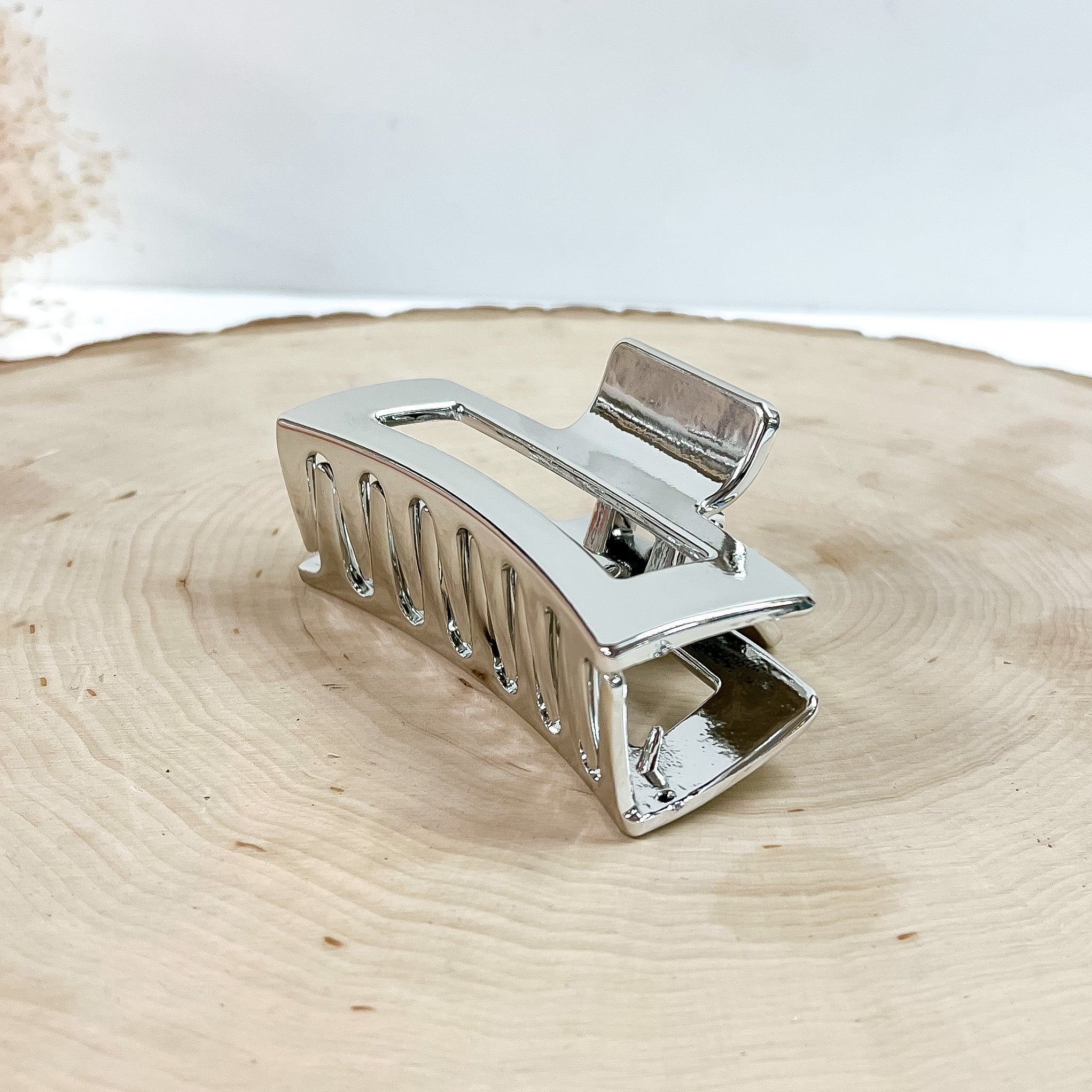 This is a rectangle metallic hair clip in silver, this clip is taken on a  slab of wood and in white background with a plant in the side as decor.