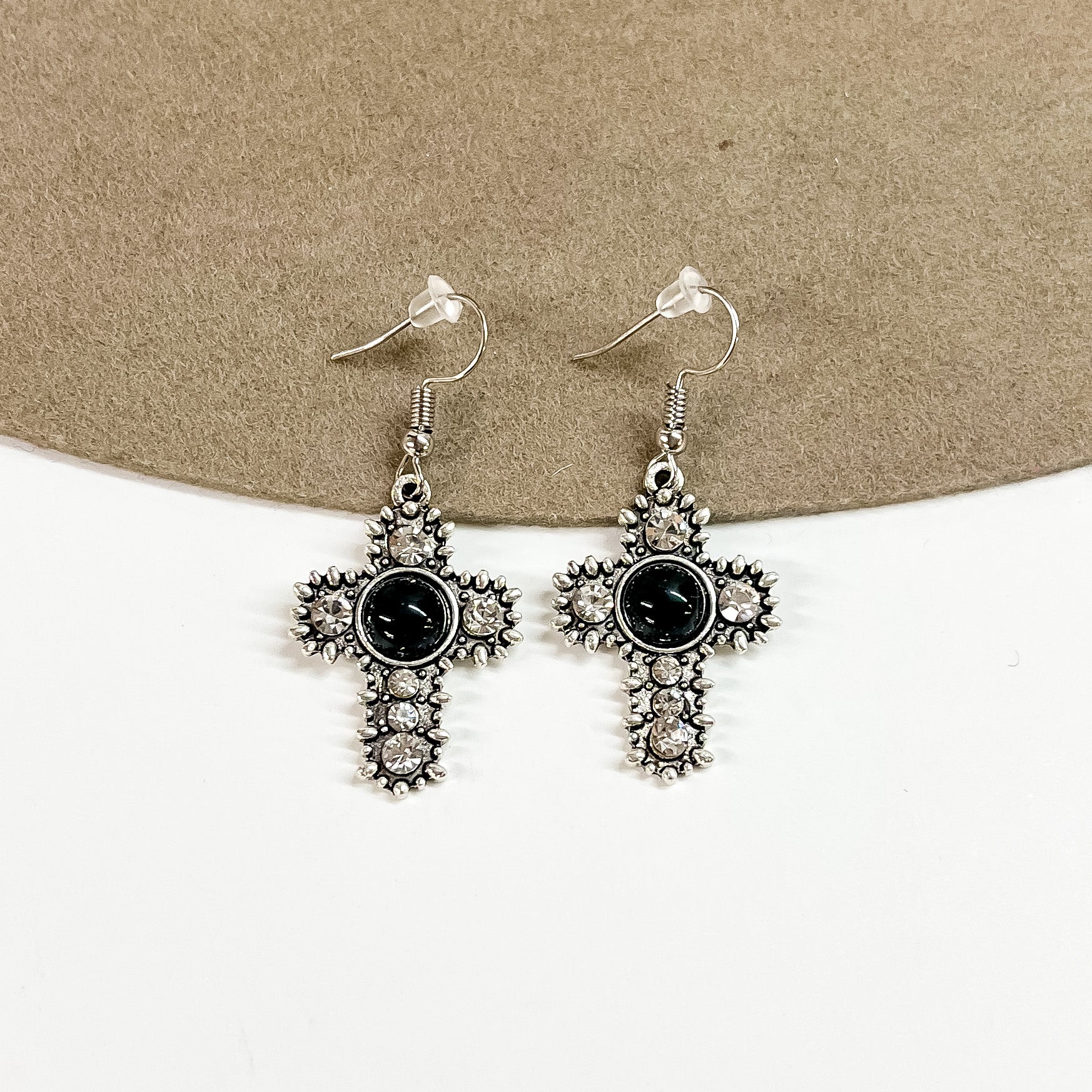 This is a pair of silver detailed cross earrings with small clear crystals  and a black circle stone in the center.  These earrings are laying on a brown felt hat brim and on a white background.