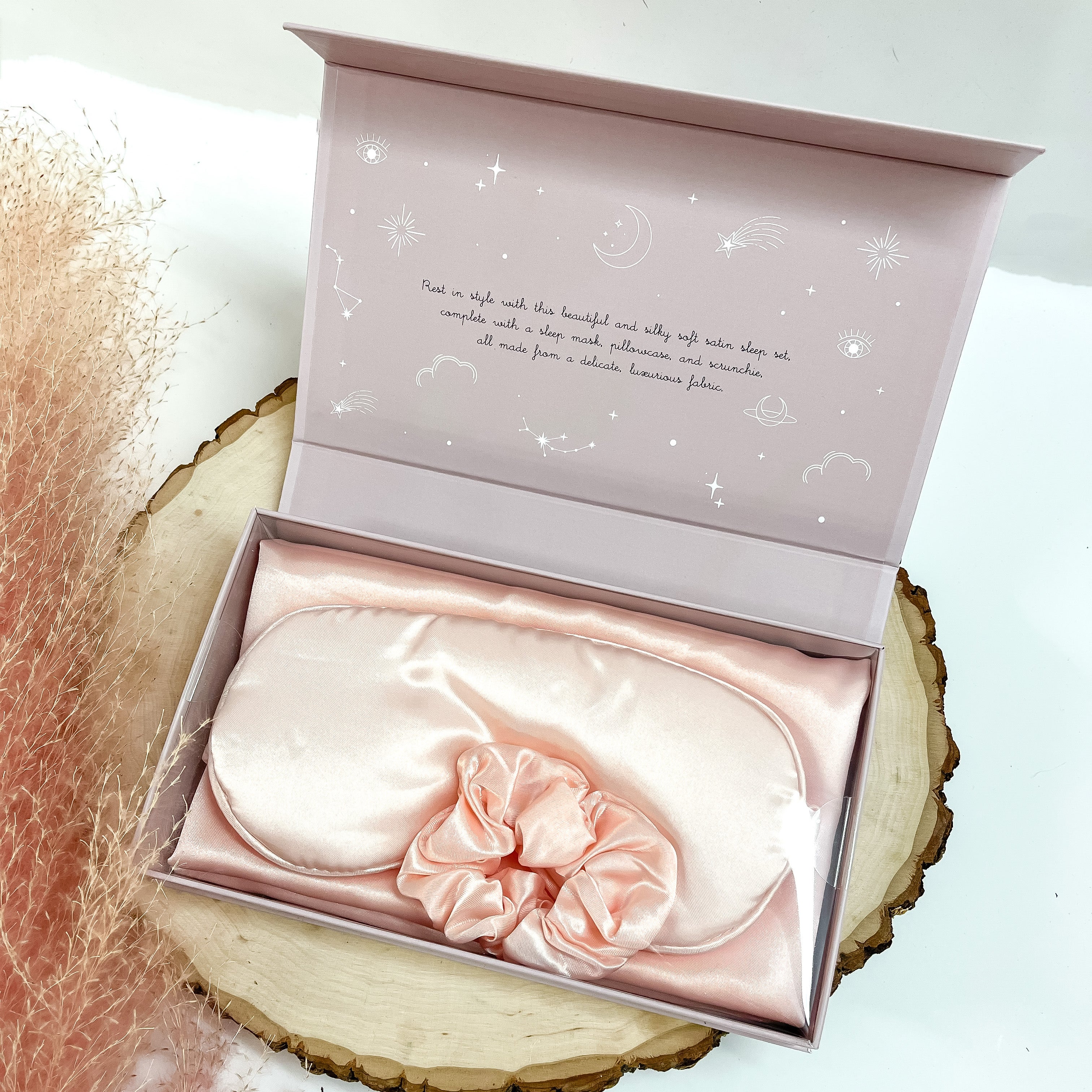 Light pink scrunchie, eye mask, and pillowcase in a light purple box. This box is pictured on a piece of wood on a white background. 
