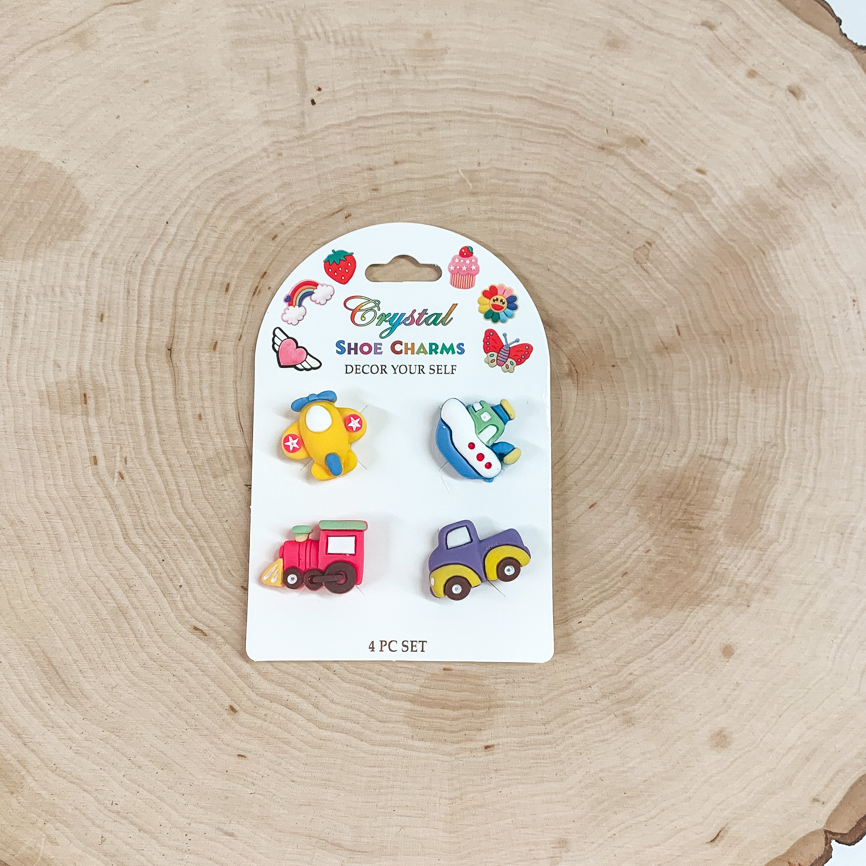 This is a pack of four small shoe charms in different colors and figures such as a plane, boat, train, and truck. These shoe charms are placed on a white card and taken on a slab of wood.