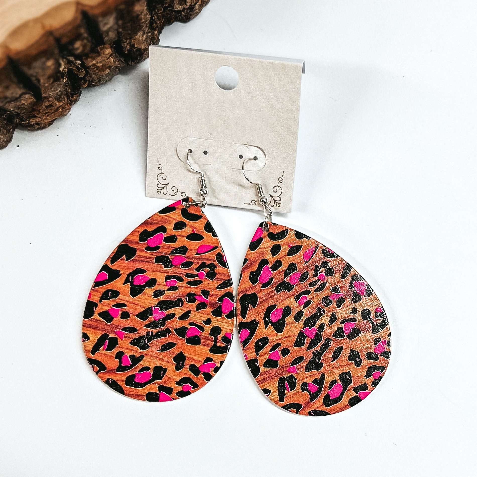 These are brown wooden teardrop earrings in leopard print and pink detailing. These earrings are placed on a white earring card holder, they are laying on a  white background with a slab of wood in the back as decor.