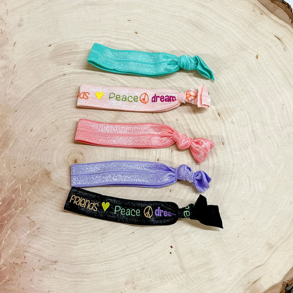 Set of five tie pony tails in different colors. Colors include, teal, light pink, pink, purple, and black. These are pictured on top of a piece of wood. 