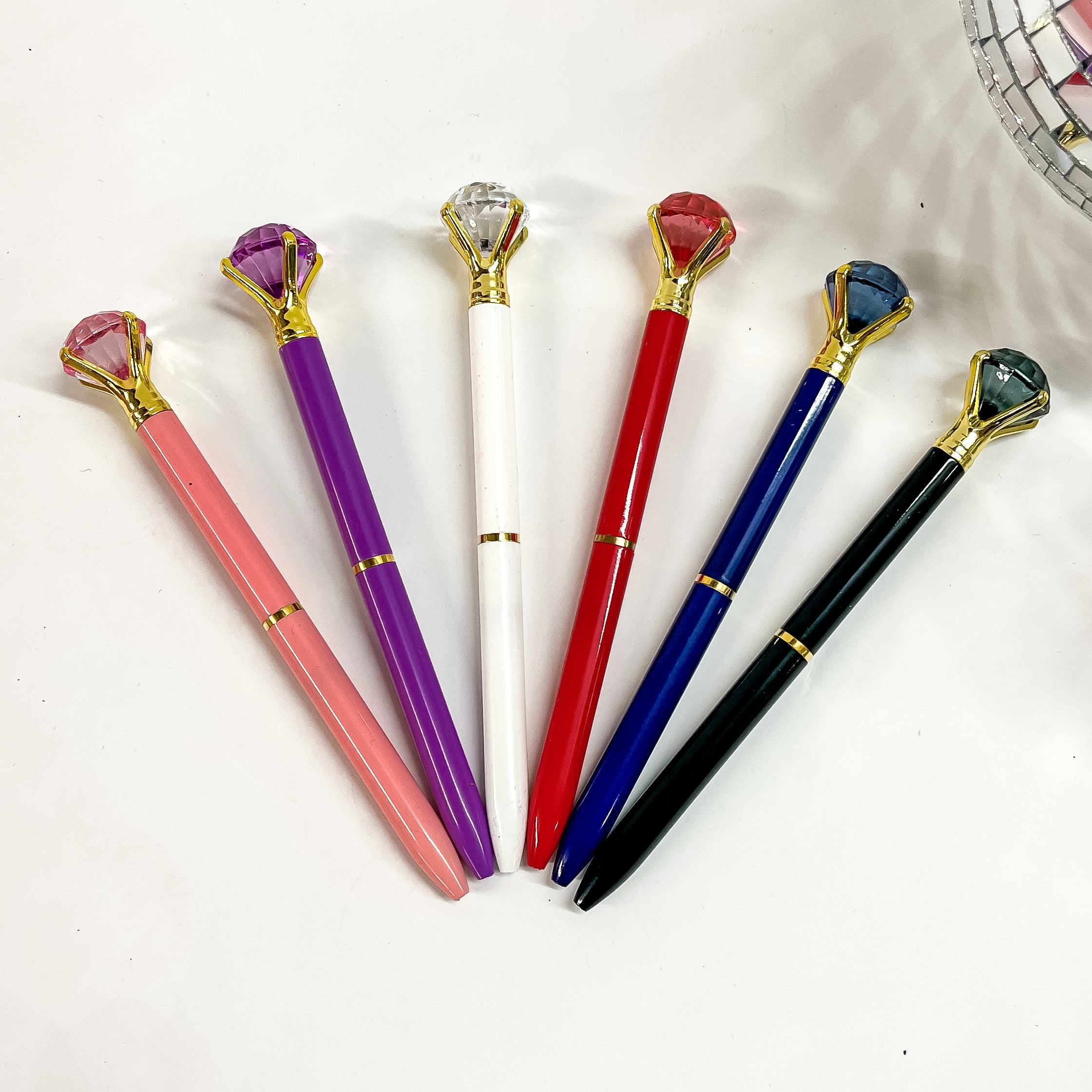 Pictured on a white background are five different colored pens. Colors include pink, purple, white, red, blue, and black. Included on each pen is a crystal on the top in the same color as the pen. 