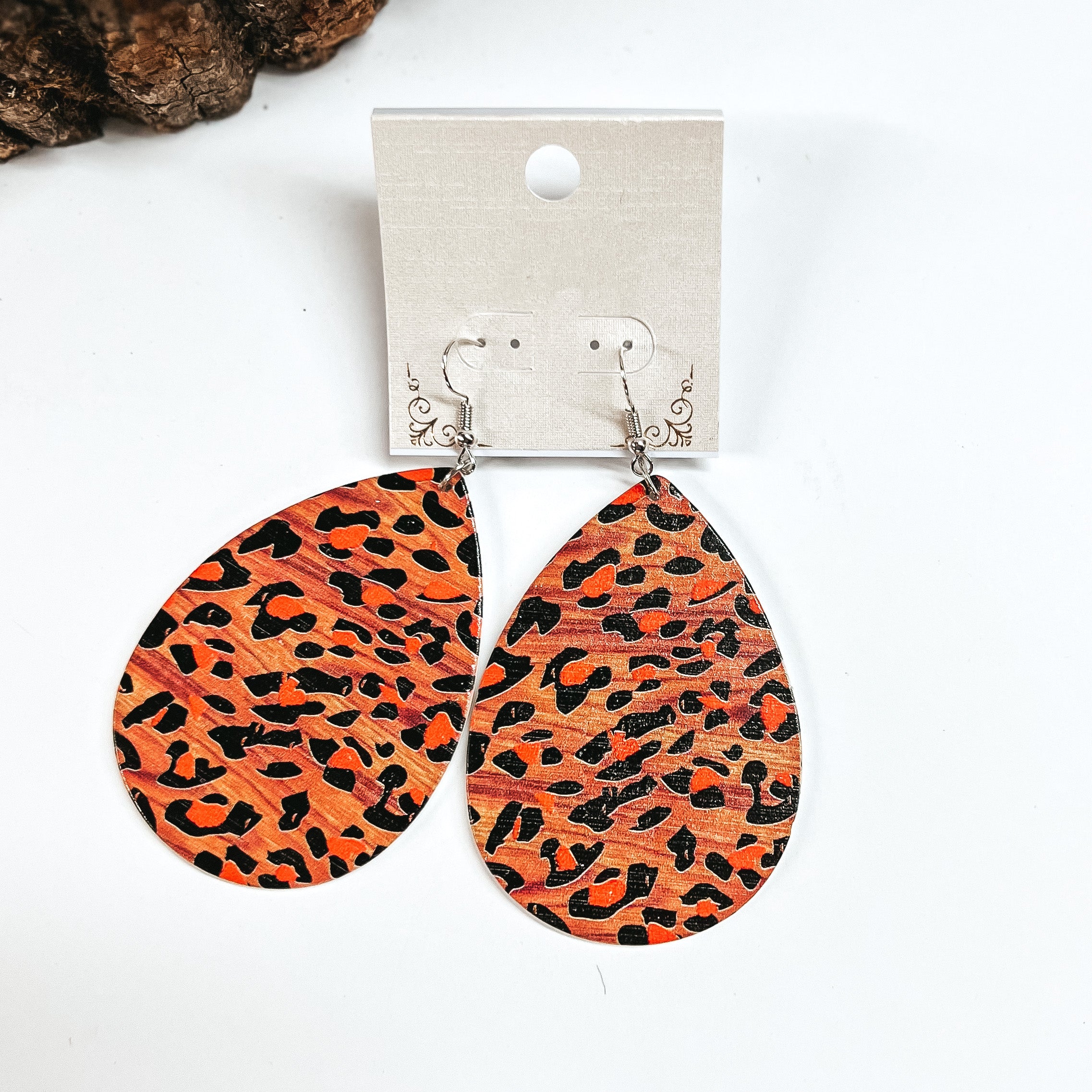 These are brown wooden teardrop earrings in leopard print and orange detailing. These earrings are placed on a white earring card holder, they are laying on a  white background with a slab of wood in the back as decor.
