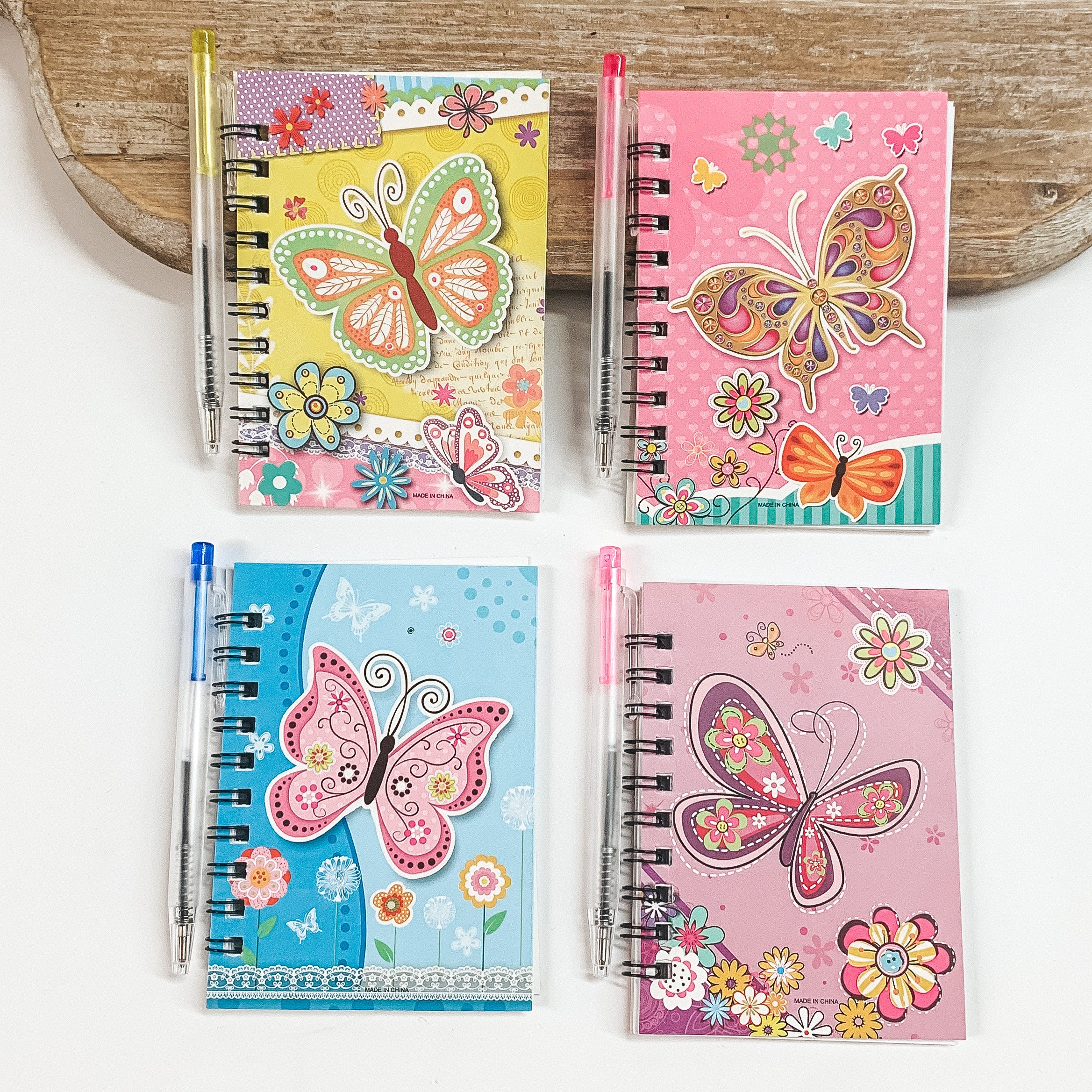 Pictured on a white background are four small notebooks with a butterfly design and a pen. Colors include yellow, pink, blue, and purple. 