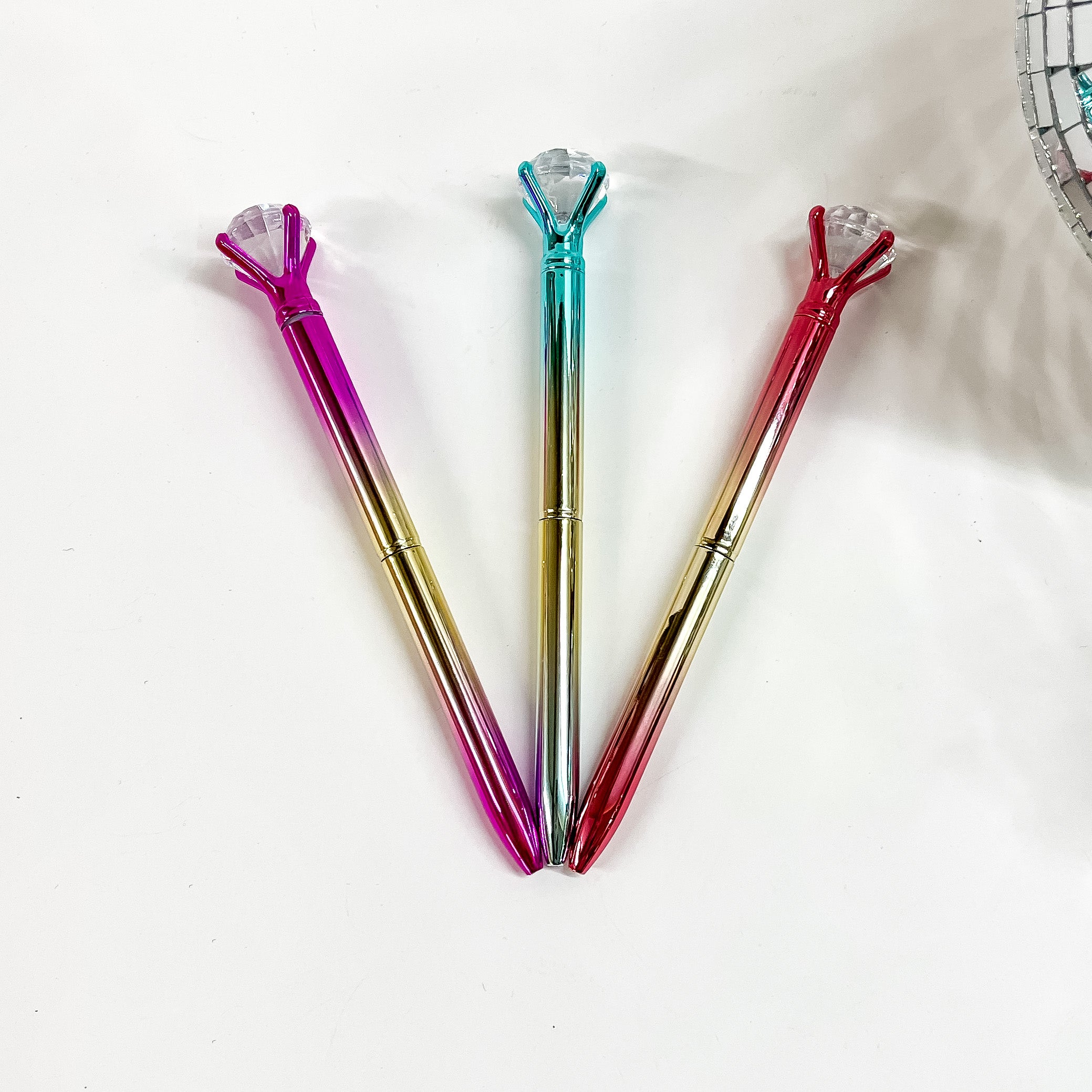 There are three metallic ombre pens with a clear diamond gem on top. From left to tight; pink/gold, turquoise/gold, and red/gold. These pens are laying on a white background with a disco ball in the side as decor.
