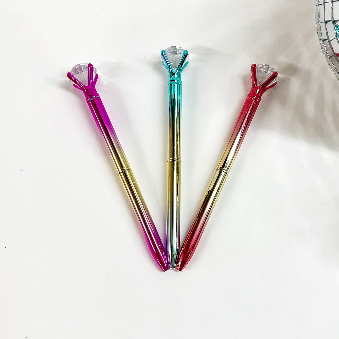 There are three metallic ombre pens with a clear diamond gem on top. From left to tight; pink/gold, turquoise/gold, and red/gold. These pens are laying on a white background with a disco ball in the side as decor.