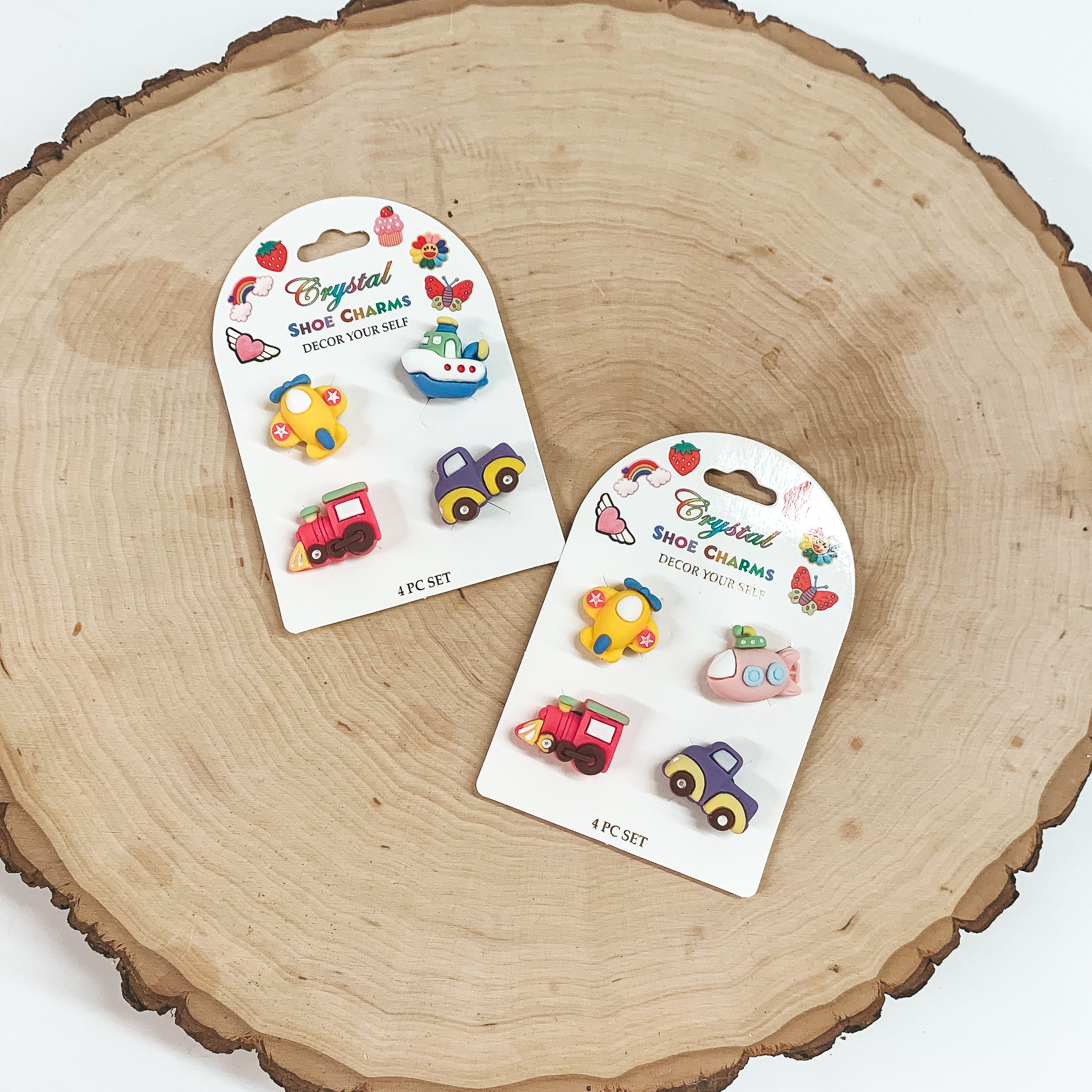 There are two packs of four small shoe charms. The left one as a boat, truck, train, and plane. The right pack has a truc, plane, train, and submarine. These shoe charms are placed on a white card and taken laying on a slab of wood and on white background.