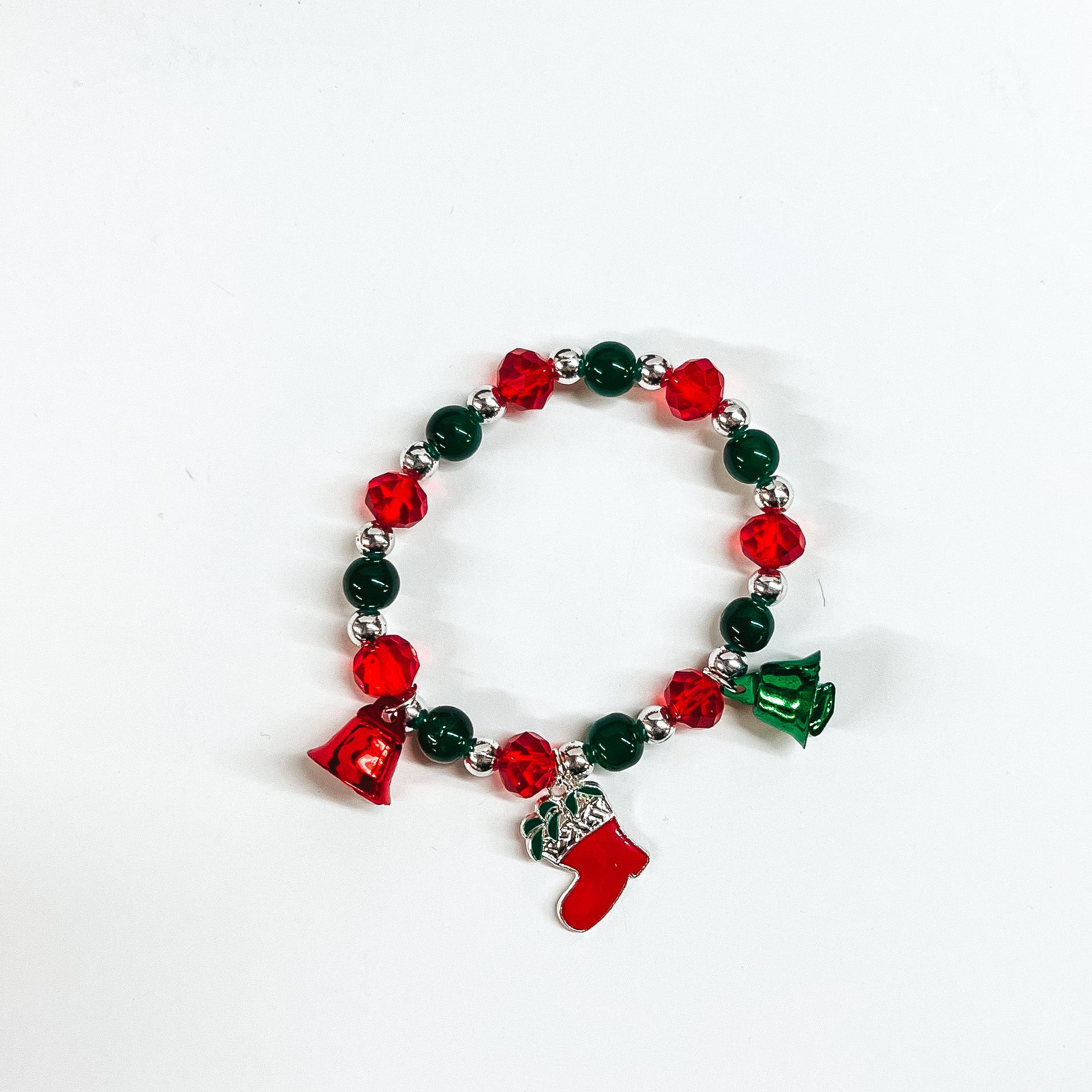 This is a beaded bracelet with three christmas charms such as two bells in red and green, and a stocking charm in red and silver. The bracelet has red crystal beads, silver circle beads, and green circle beads. This bracelet is laying on a white background.