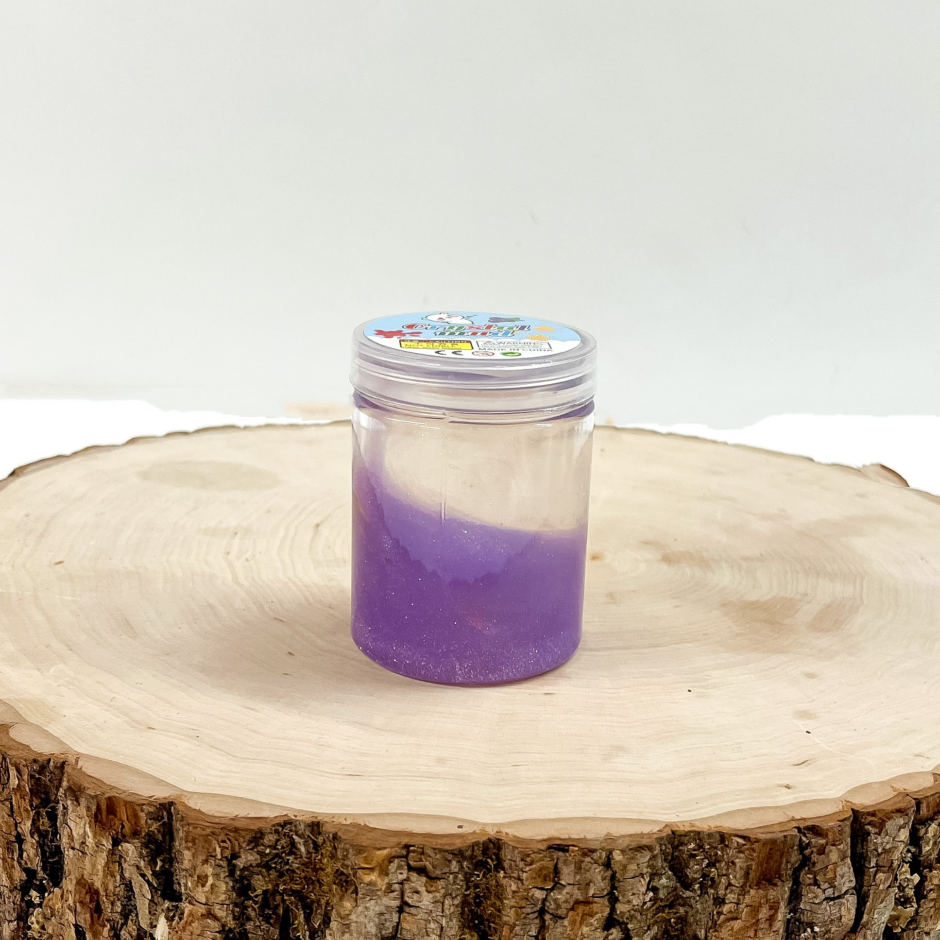 This is a clear slime with purple glitter, this container is taken on a slab of  wood and on a white background.