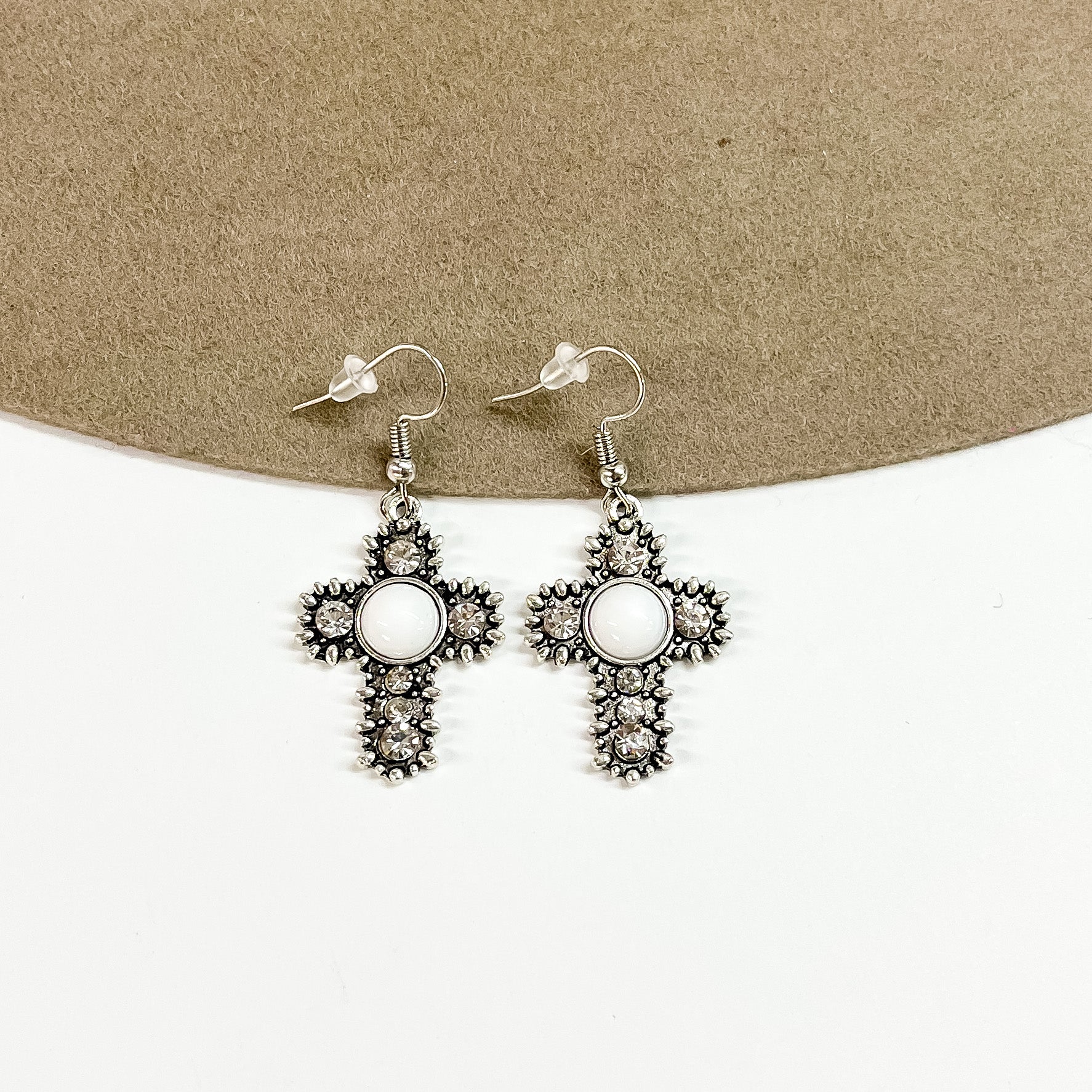 This is a pair of silver detailed cross earrings with small clear crystals and a white circle stone in the center. These earrings are laying on a brown felt hat brim and on a white background.