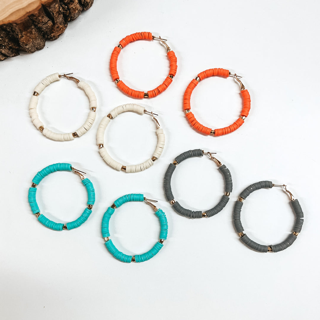 There are five pairs of beaded hoop earrings in different colors. From top to bottom; coral, ivory, gray, and turquoise. All hoops have disc beads and gold spacers. These earrings are taken on a white background with a slab of wood in the back as decor.