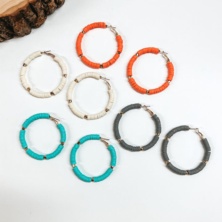 There are five pairs of beaded hoop earrings in different colors. From top to bottom; coral, ivory, gray, and turquoise. All hoops have disc beads and gold spacers. These earrings are taken on a white background with a slab of wood in the back as decor.