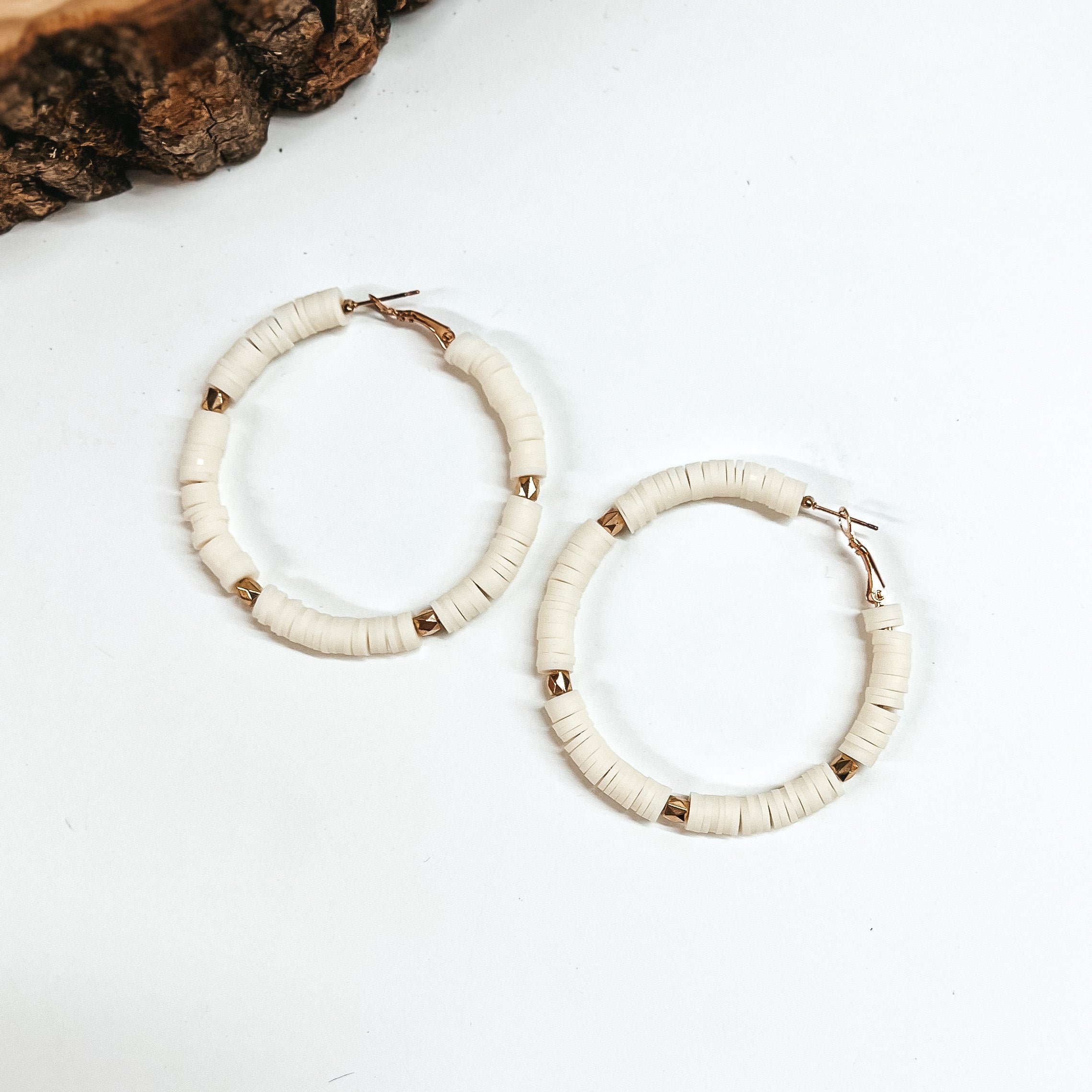 This is a pair of ivory disc beaded hoop earrings with gold spacers. These  earrings are taken on a white background with a slab of wood in the back  as decor.