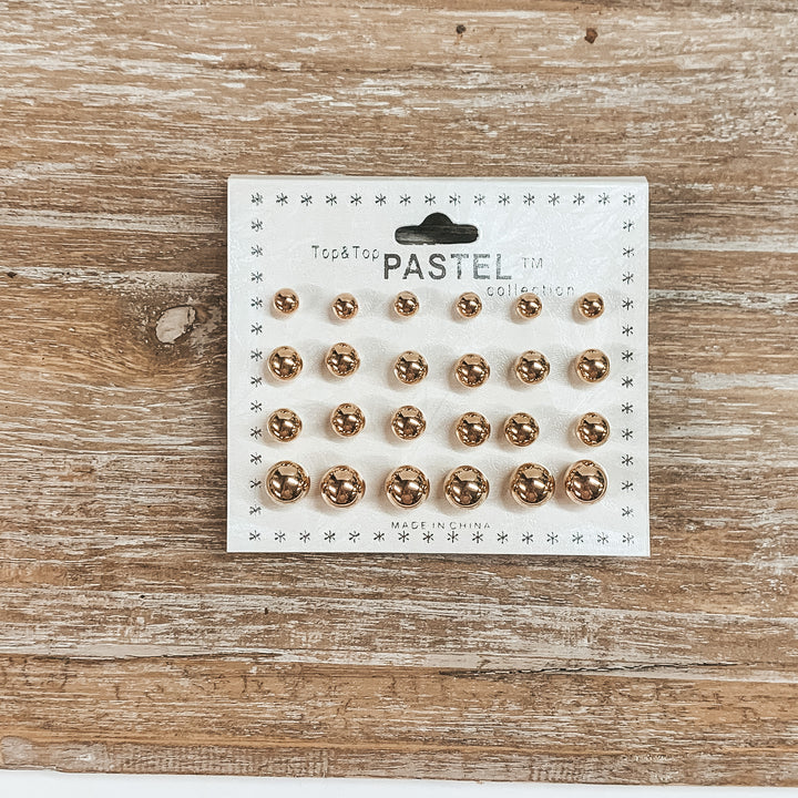 This is a pack of twelve gold circle stud earrings in different sizes. These  earrings are placed on a white earring card holder and laying on a wooden slate.