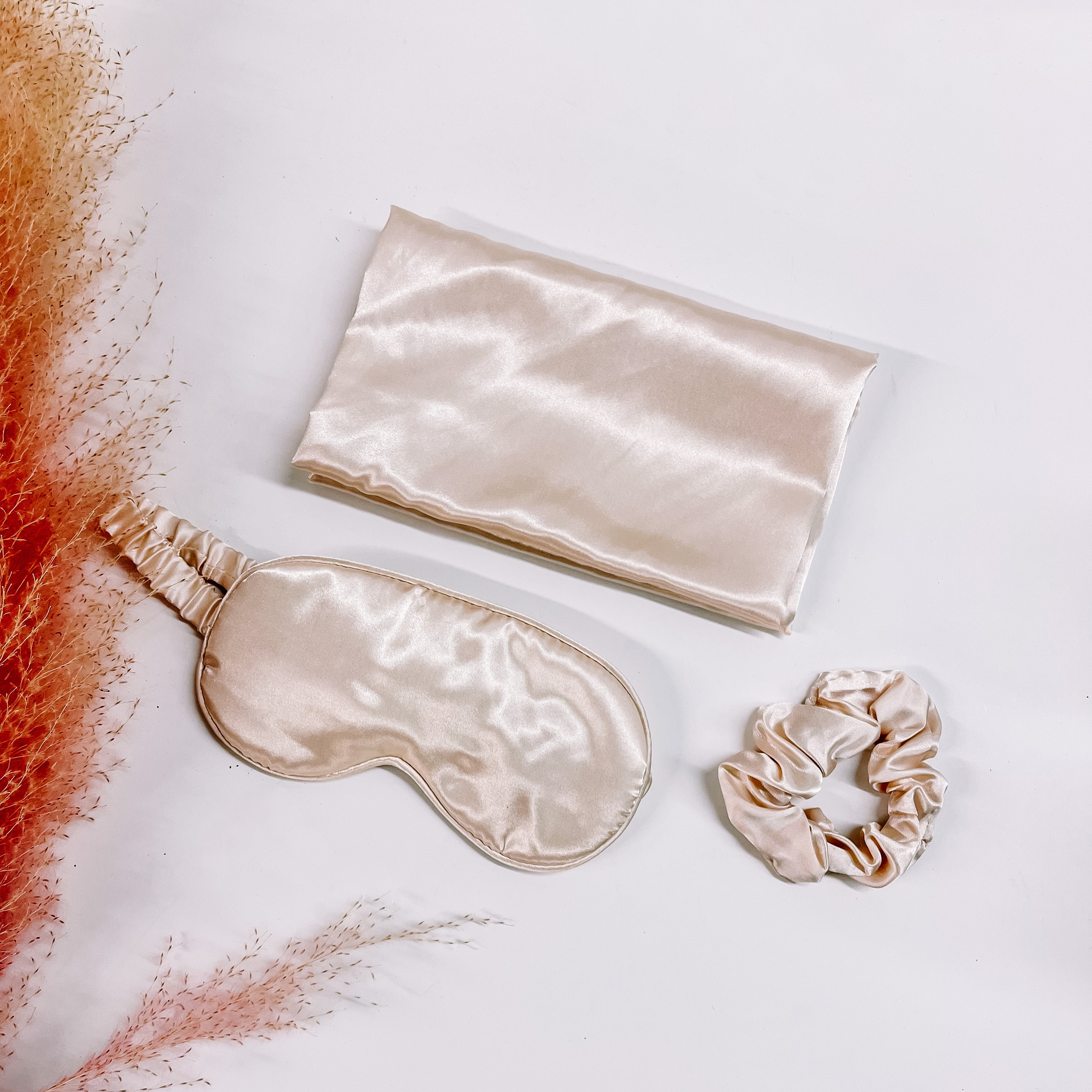This is a satin pillow case, eye mask, and srunchie in invory. These items are taken on a white background with a pink plant in the side as decor.