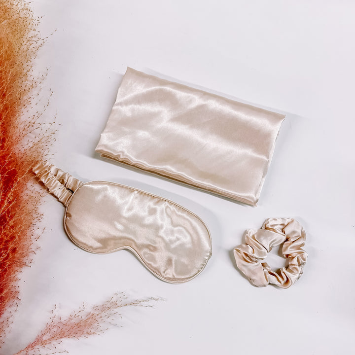This is a satin pillow case, eye mask, and srunchie in invory. These items are taken on a white background with a pink plant in the side as decor.