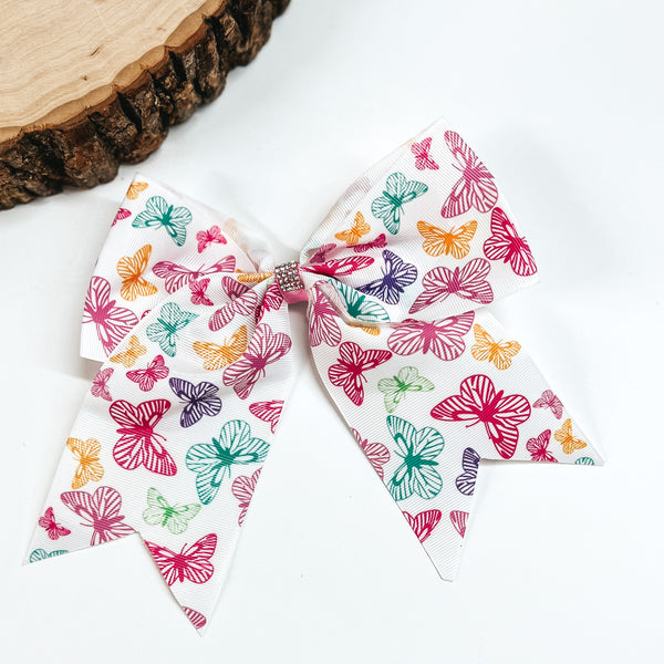 White colored hair bow with a multicolored butterfly print. It also includes a clear crystal center. This bow is pictured on a white background. 