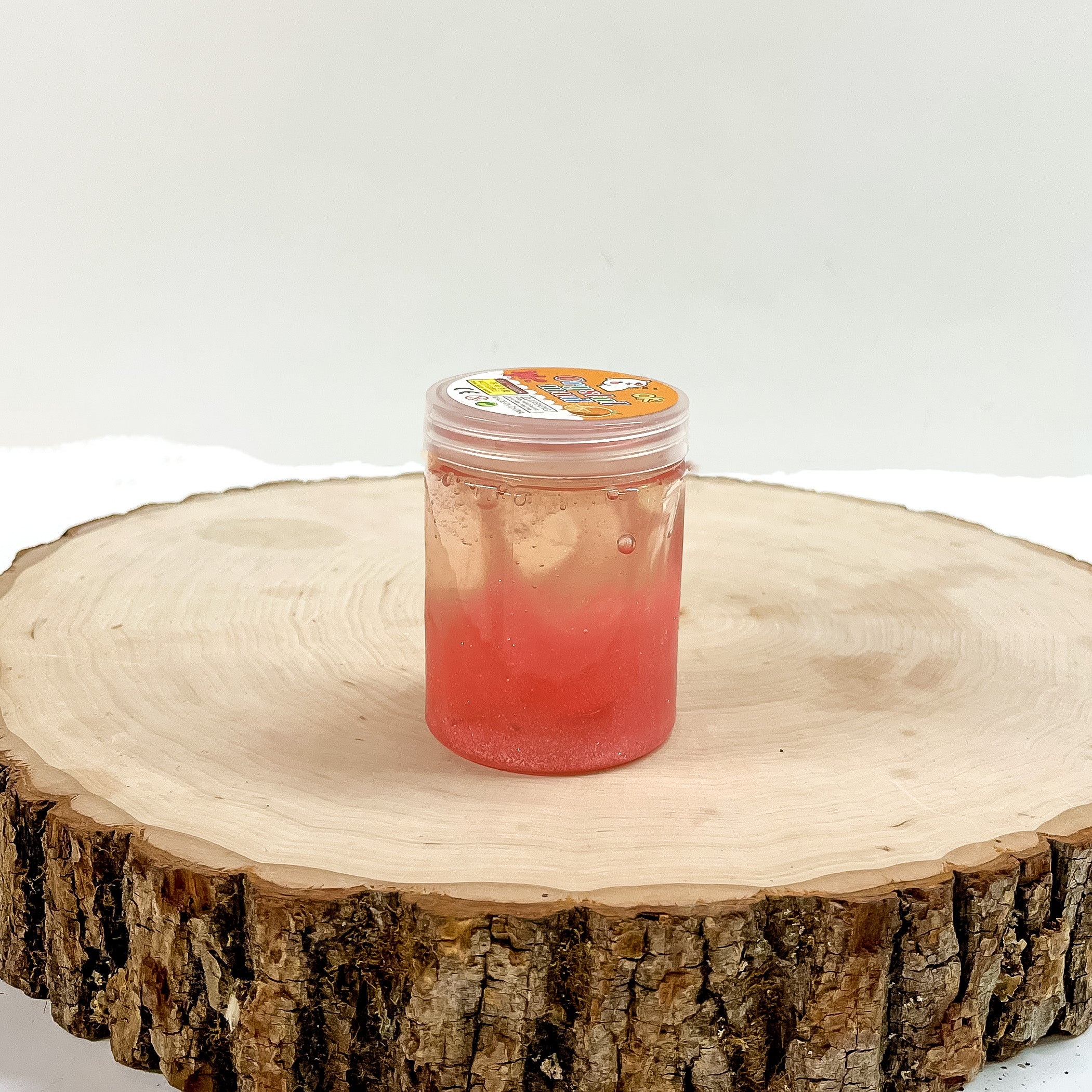 This is a clear slime with coral glitter, this container is taken on a slab of  wood and on a white background.