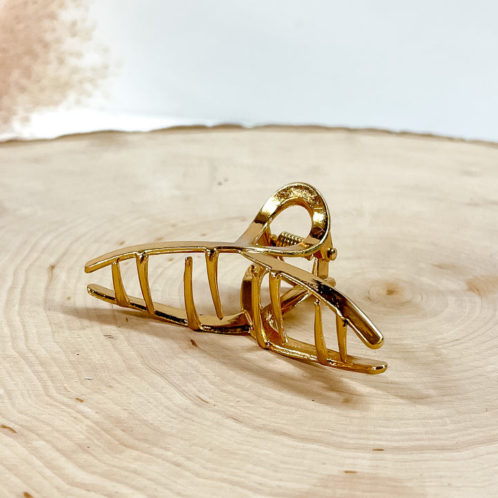 This is how the inside of the clip looks, small thin grabbers. This is a small, gold, smooth, hair clip laying on a slab of wood with a pink plant in the side as decor.