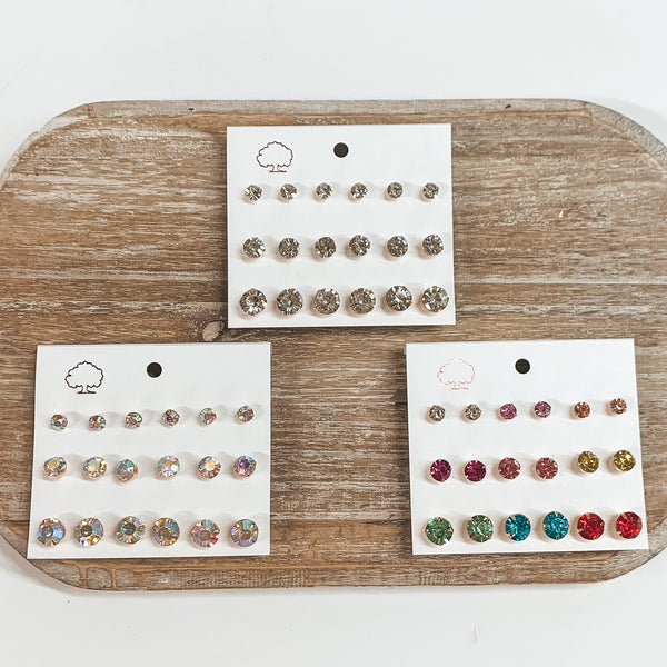 There are a pack of three earring sets of nine. The left one has different sizes od ab crystals studs, the middle one has clear crystal studs in a gold setting, and the right has multicolored crystals. These earrings are taken on a white earring card holder and they are laying on a wooden slate and on a white background.