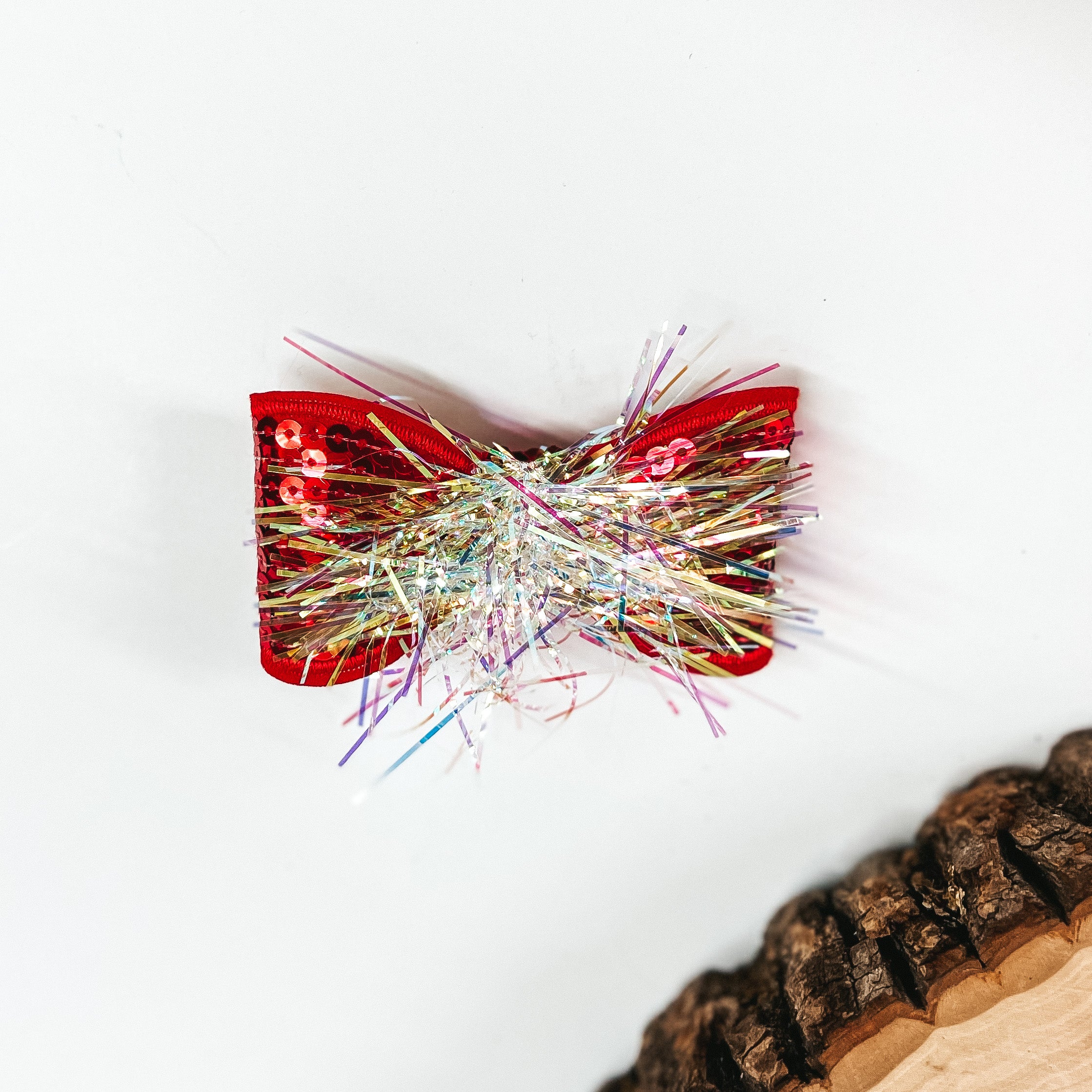 This is a small red sequin bow with a tinsel center, this bow is taken on a  white background with a slab of wood in the corner as decor.