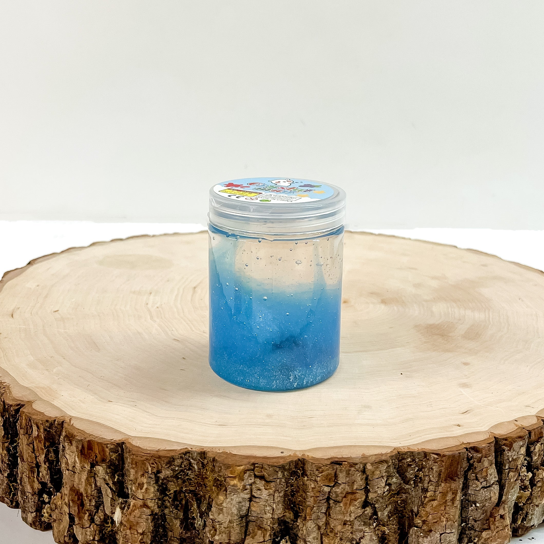 This is a clear slime with blue glitter, this container is taken on a slab of  wood and on a white background.