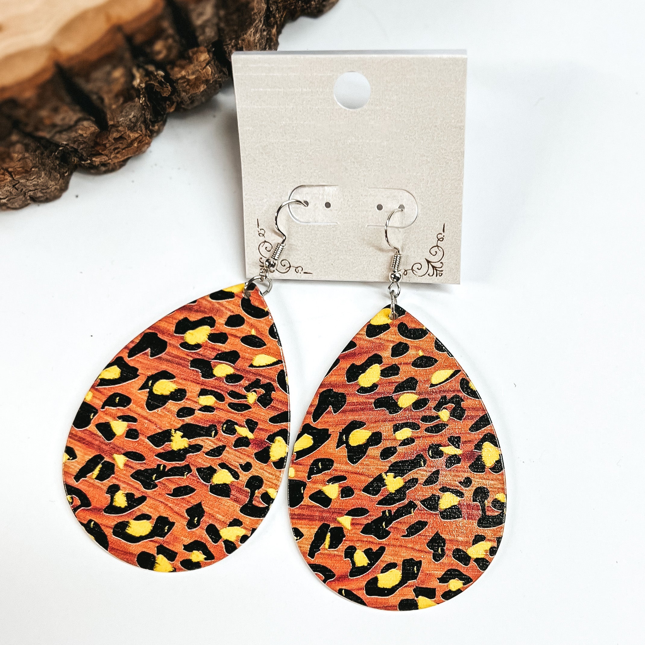 These are brown wooden teardrop earrings in leopard print and yellow detailing. These earrings are placed on a white earring card holder, they are laying on a  white background with a slab of wood in the back as decor.
