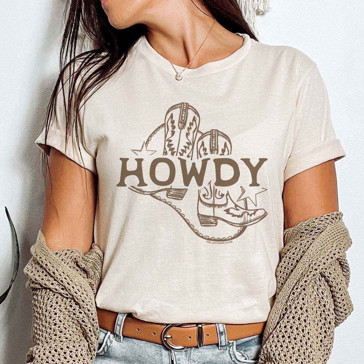 A cream colored short sleeve shirt with cuffed sleeves featuring a graphic of a pair of brown cowboy boots with the word "Howdy" running through the middle. There are brown stars on both the start and end of the text with an unsymmetrical line border going around the graphic. Item is pictured on a plain white background.