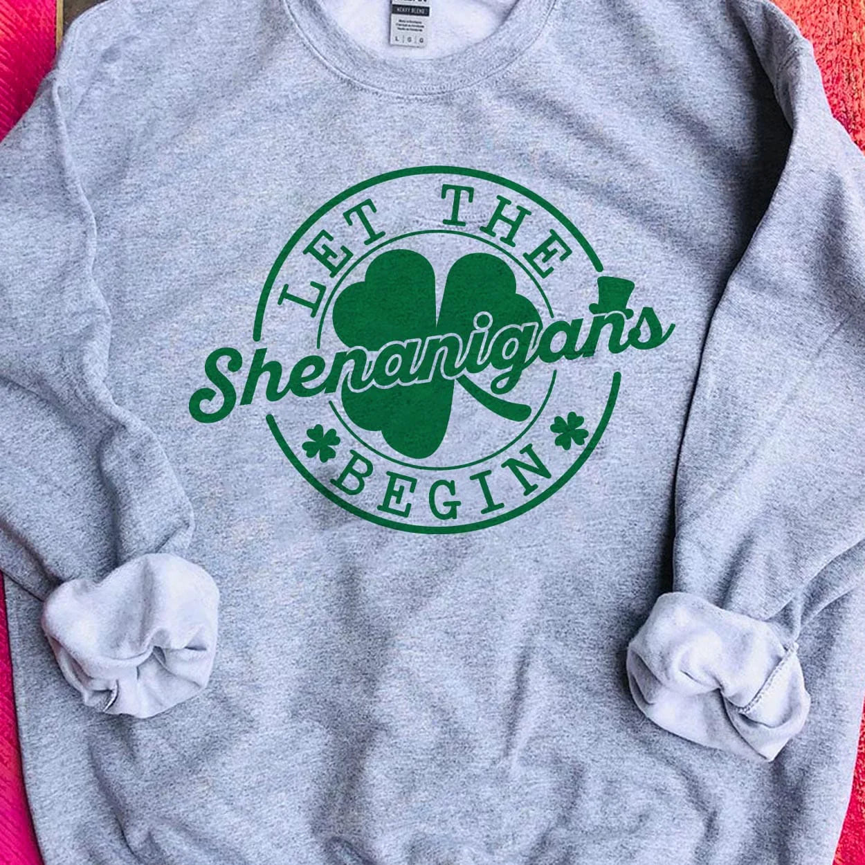 A gray crew neck sweatshirt featuring a large circular graphic saying "Let the shenanigans begin" in dark green with a clover behind "shenanigans" and little clovers before and after "begin". Item is pictured on a pink background