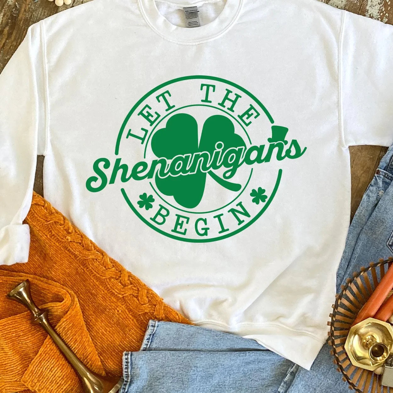 A white short sleeve tee featuring a large circular graphic saying "Let the shenanigans begin" in dark green with a clover behind "shenanigans" and little clovers before and after "begin". Item is pictured on a brown background