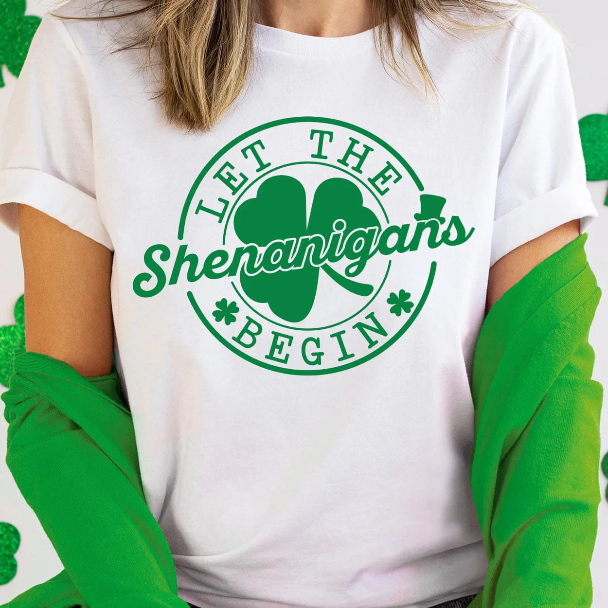 A white short sleeve tee featuring a large circular graphic saying "Let the shenanigans begin" in dark green with a clover behind "shenanigans" and little clovers before and after "begin". Item is pictured on a white and green background