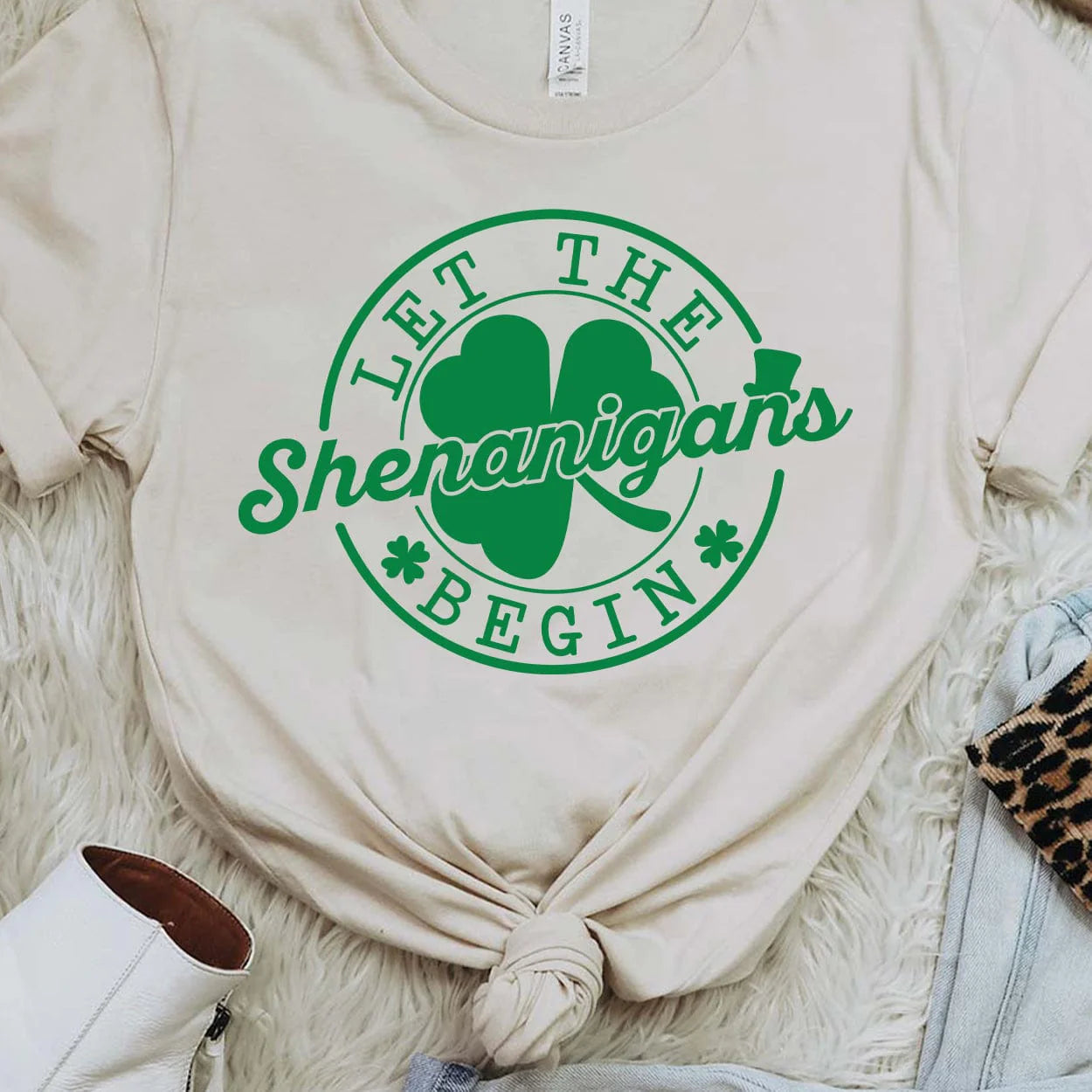 A cream short sleeve tee featuring a large circular graphic saying "Let the shenanigans begin" in dark green with a clover behind "shenanigans" and little clovers before and after "begin". Item is pictured on a white background
