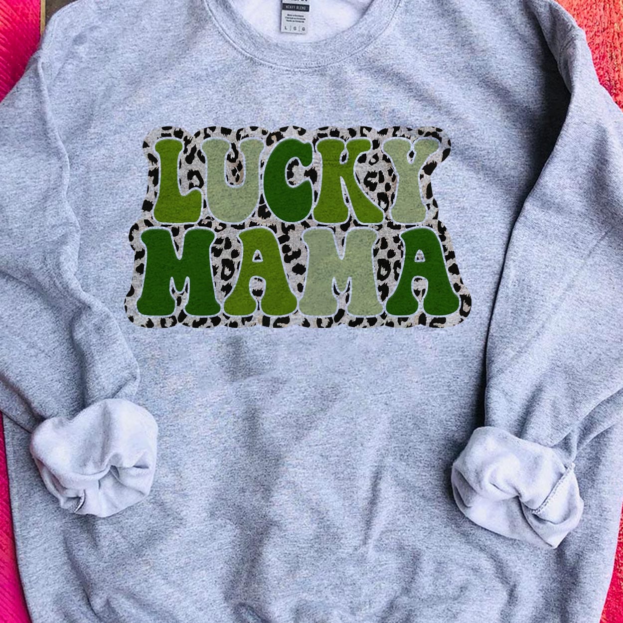 A gray crew neck sweatshirt featuring the words "Lucky mama" in various shades of green with a small leopard border around the text. Item is pictured on a pink background.