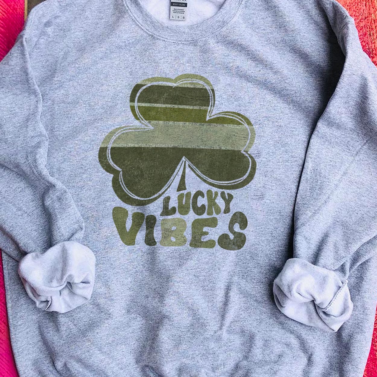 A gray crew neck sweatshirt featuring a large graphic of a clover with various shades of green and the words "lucky vibes" below it with each letter coordinating to a color in the clover. Item is pictured on a pink background.