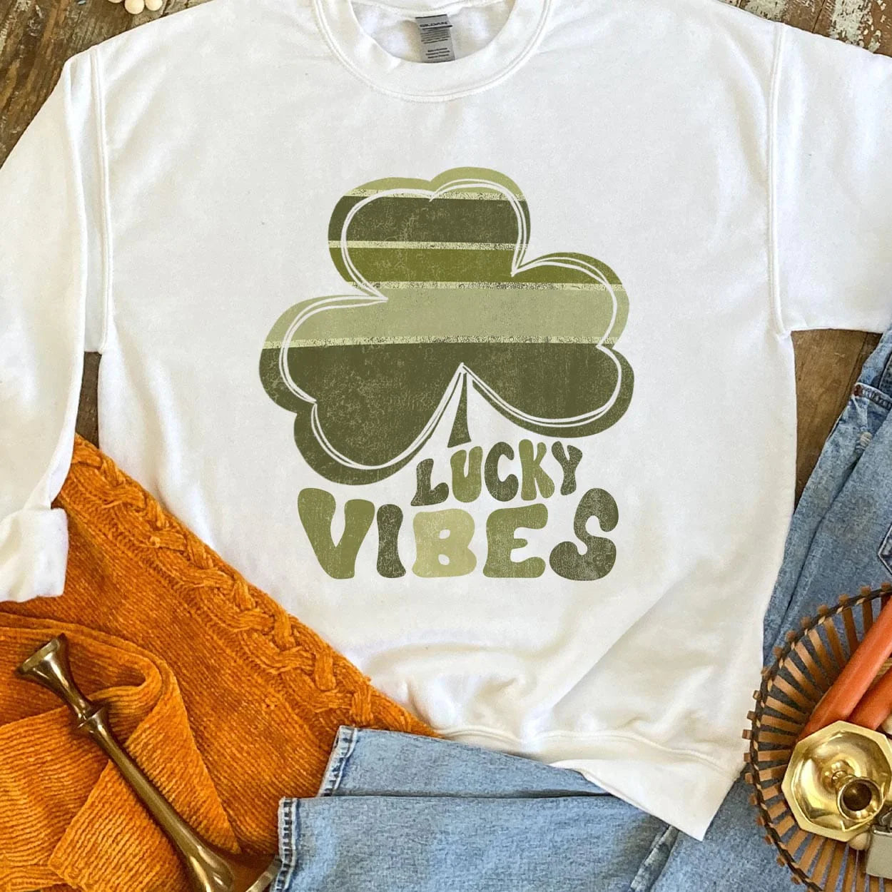 A white crew neck sweatshirt featuring a large graphic of a clover with various shades of green and the words "lucky vibes" below it with each letter coordinating to a color in the clover. Item is pictured on a brown background.