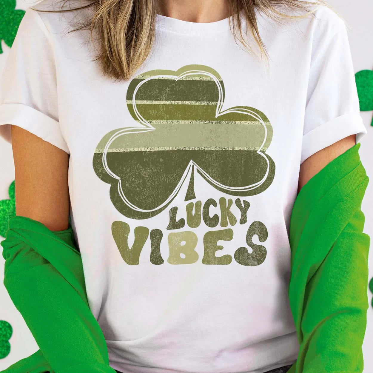 A white short sleeve tee with cuffed sleeves featuring a large graphic of a clover with various shades of green and the words "lucky vibes" below it with each letter coordinating to a color in the clover. Item is pictured on a white and green background.