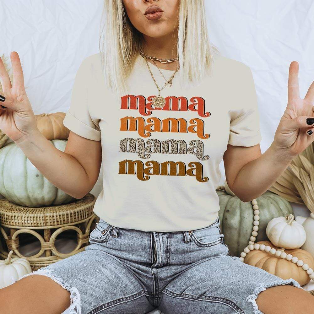 A short sleeve cream color crew neck tee with folded sleeves and the word "mama" in a stack of four. The top text is in red, the second is orange, the third is leopard print, and the bottom is brown. Item is pictured on a white background with pale colored pumpkins. 