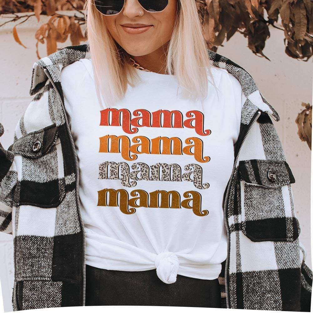 A short sleeve white crew neck tee with folded sleeves and the word "mama" in a stack of four. The top text is in red, the second is orange, the third is leopard print, and the bottom is brown. Item is pictured on a white background with brown leaves.