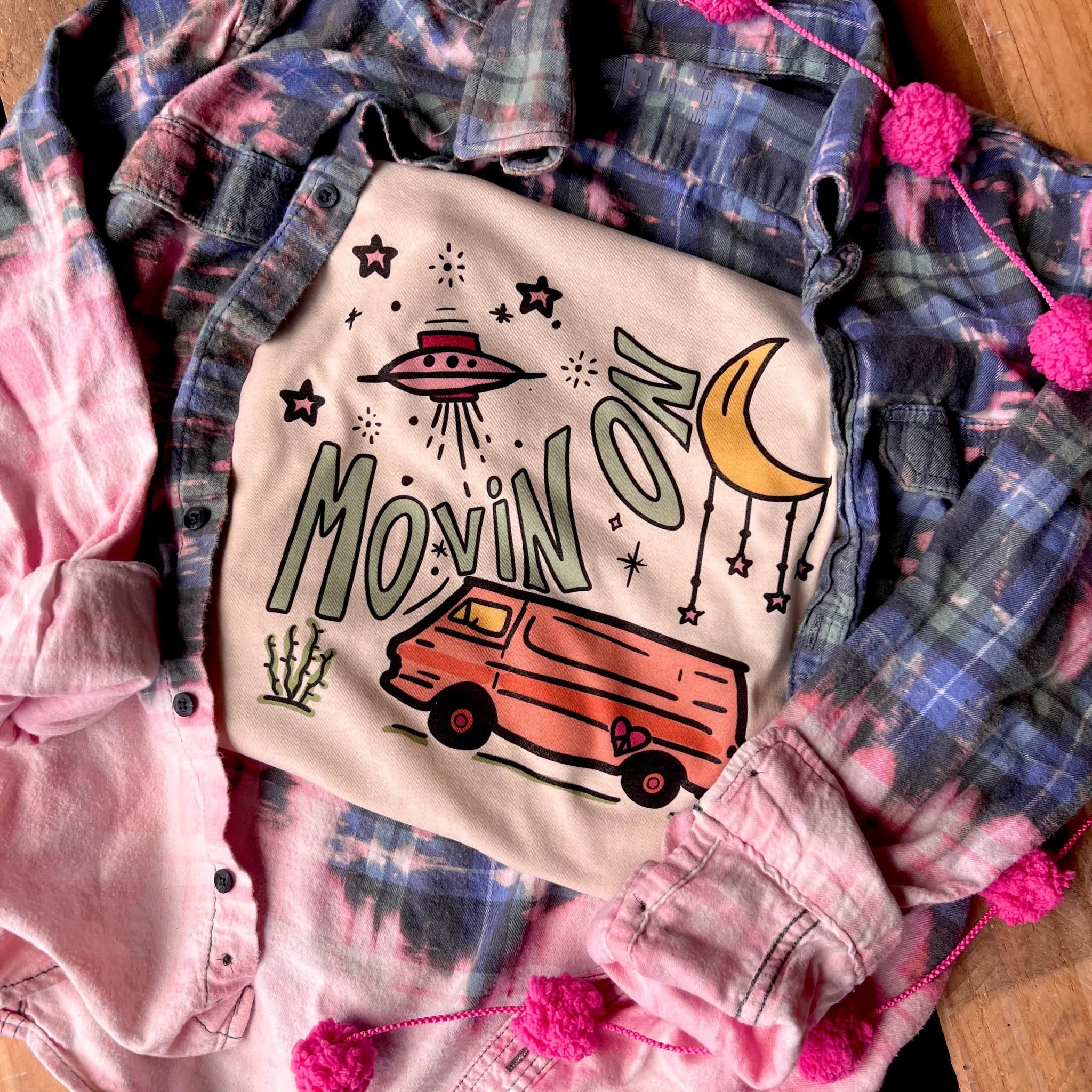 This cream graphic tee includes a crew neckline, short sleeves, and cute hand drawn design of a spaceship, camper van, moon with stars hanging from it, and the words "MOVIN ON" in a light turquoise between the spaceship and van. This tee is shown in this photo as a folded flat lay on top of a bleached button up plaid top. 