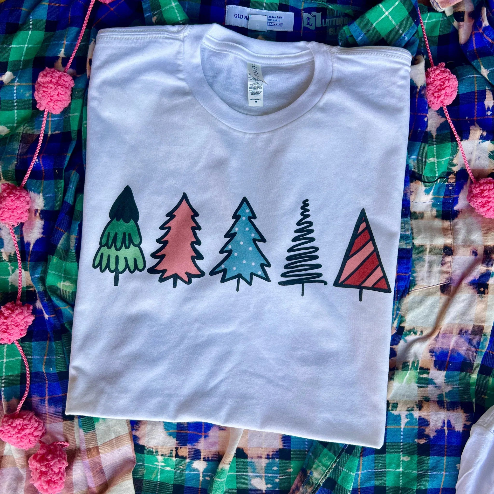 This white tee includes a crew neckline, short sleeves, and a cute Christmas graphic of five Christmas trees with different colors and prints. This is shown here as a folded flat lay. 