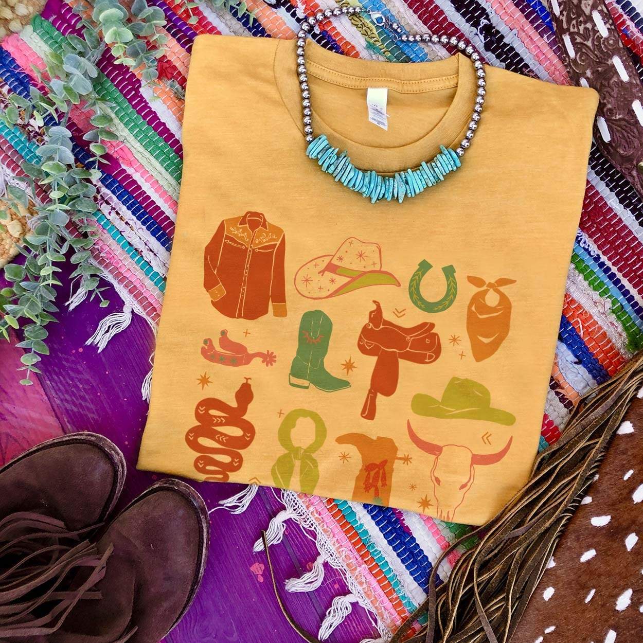 This mustard yellow graphic tee has a variety of cowgirl necessities and western finds. Items include a cowboy hat, a snake, a wild rag, and many more in various pops of colors. This is a Bella + Canvas tee with short sleeves and a crew neckline. It is shown here styled as a flat lay with a turquoise necklace and brown booties. 