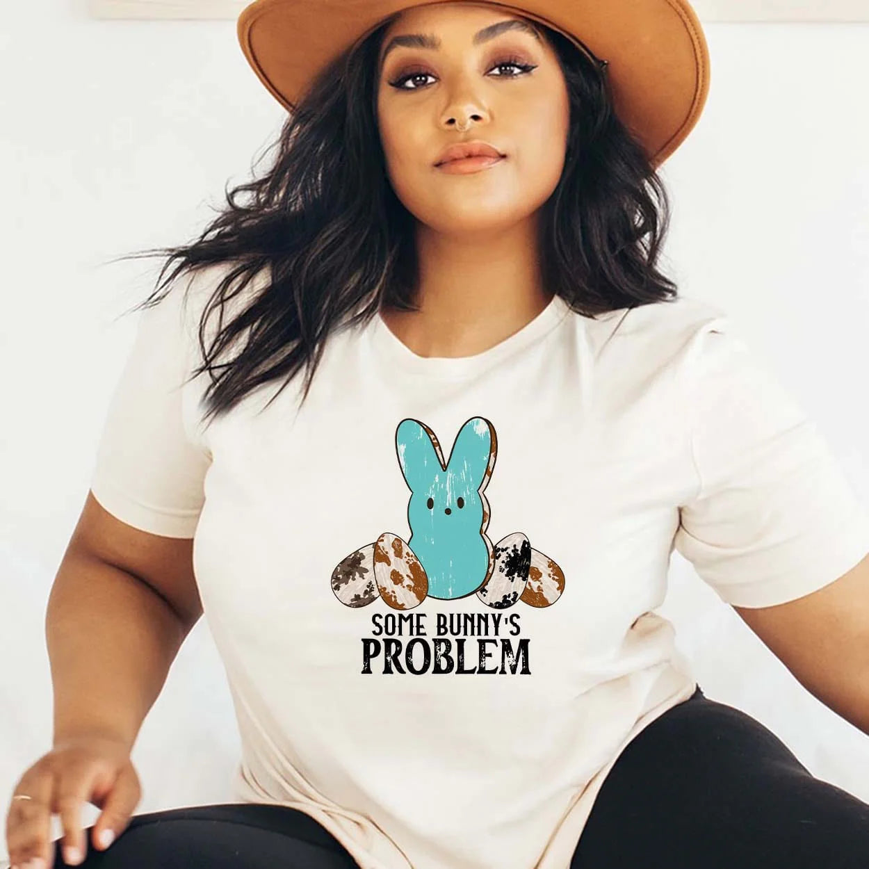 A cream colored short sleeve crew neck tee with a graphic of a blue Peep with a black and blue cow print egg on both sides with the text "Some Bunny's problem" Item is pictured on a plain white background