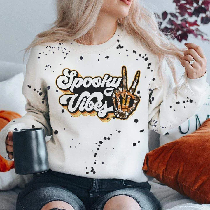 model is wearing a white, splatter paint graphic sweaterhsirt that reads "spooky vibes" in a groovy font with a skeleton hand doing a peace sign hand gesture. Model is sitting in front of a fall background and has it paired with black, ripped jeans. 